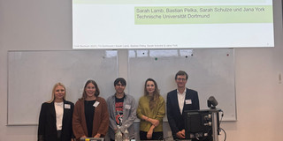 Drei Frauen und zwei Männer stehen hinter einem Hörsaalpult und schauen in Richtung Kamera.