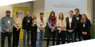 Gruppenfoto von zehn Personen, die erfreut in Richtung Kamera schauen. Im Hintergrund ist eine Powerpoint-Präsentation zum Thema "Berufliche Übergänge und Teilhabe an Arbeit unter Inklusionsverzeichen" zu sehen.