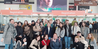 Gruppenfoto mit vielen Personen vor dem Eingang einer Messe. Im Hintergrund sind große Bildschirme mit Veranstaltungshinweisen zu sehen.